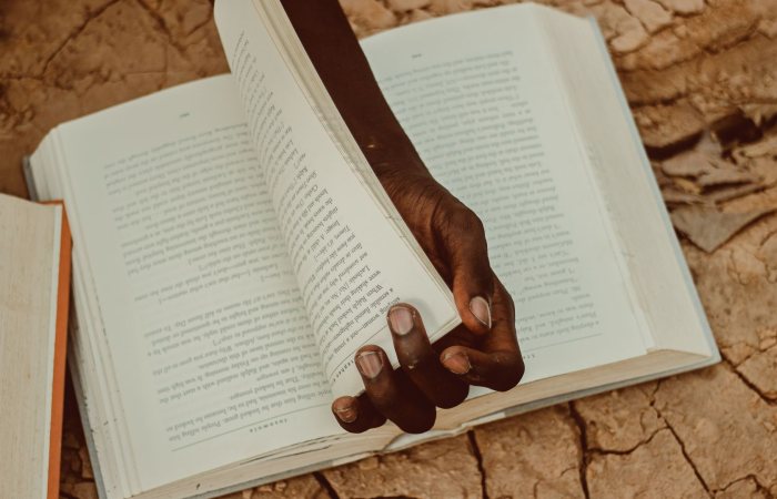 A person holds a book open on parched, cracked earth, symbolizing knowledge amidst adversity.